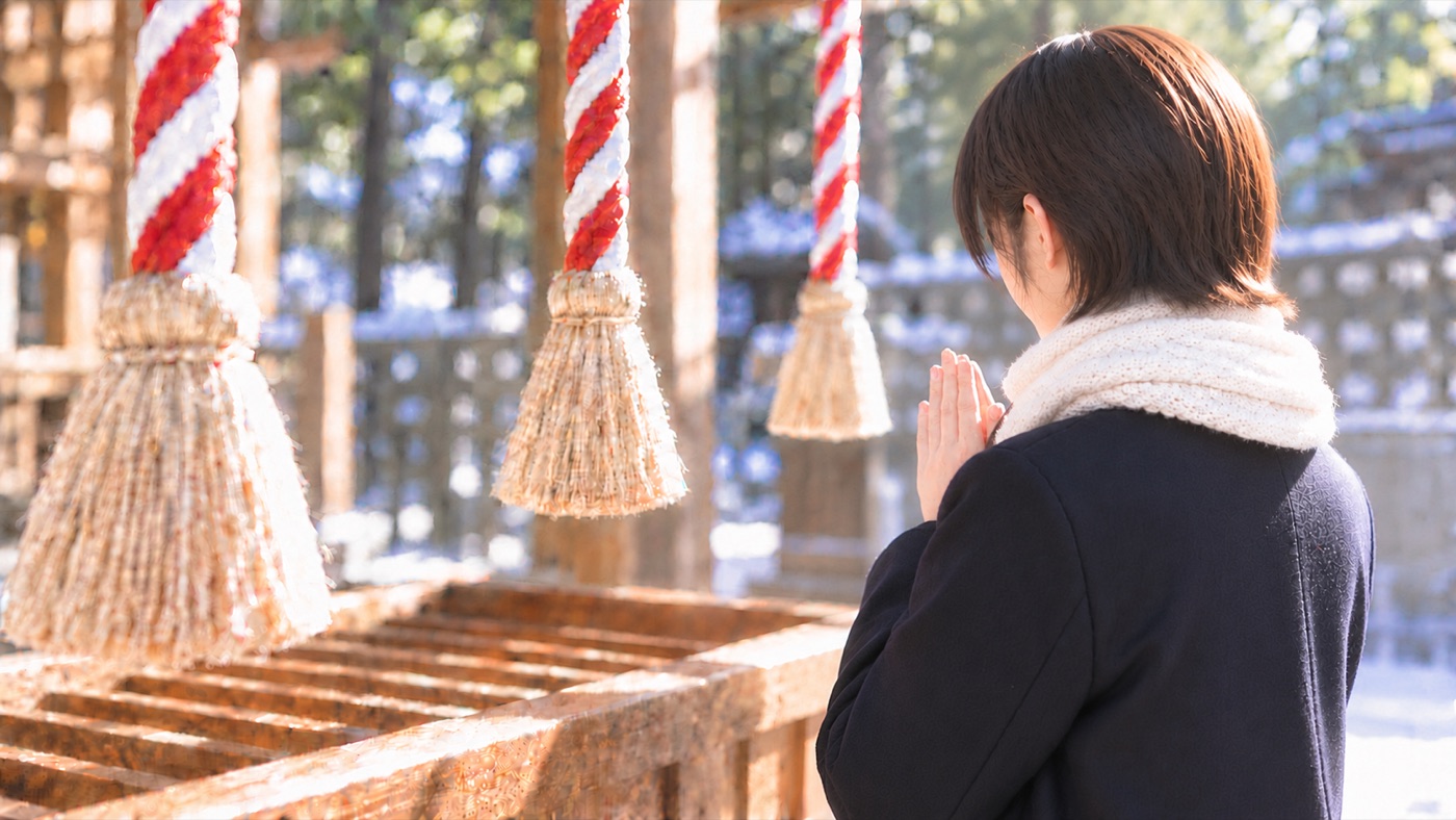 雪の残る神社で、紅白の鈴緒の前に立ち手を合わせる学生の後ろ姿